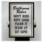 Person holding a large black framed Bathroom Rules humorous quote canvas art print showing print size scale and bold vintage distressed typography with Don't Miss Wipe Good Flush It Wash Up Git Gone text detail